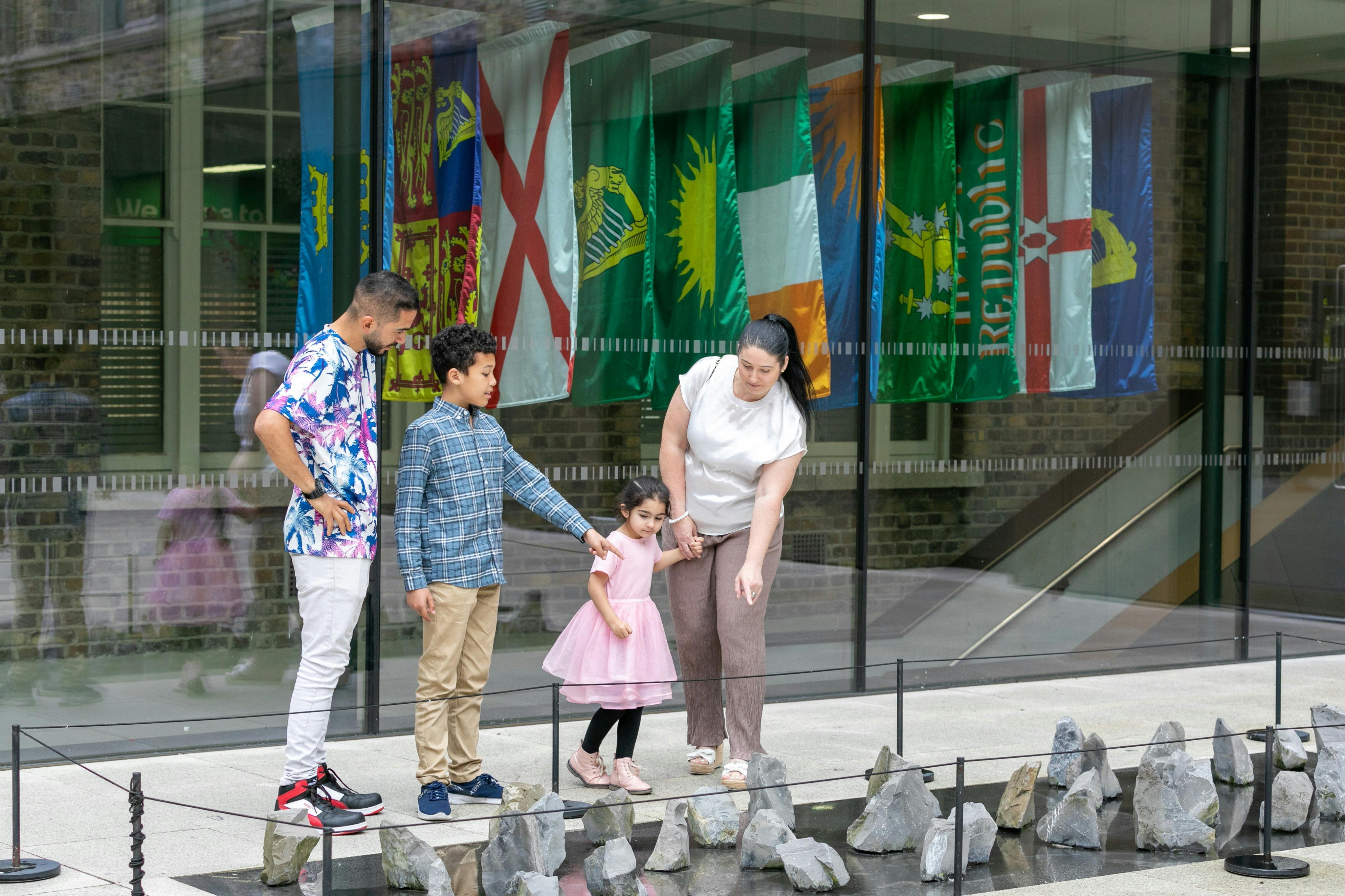 Eine vierköpfige Familie, darunter zwei Kinder, betrachtet eine Felsenausstellung vor einem Gebäude mit verschiedenen Nationalflaggen im Hintergrund.