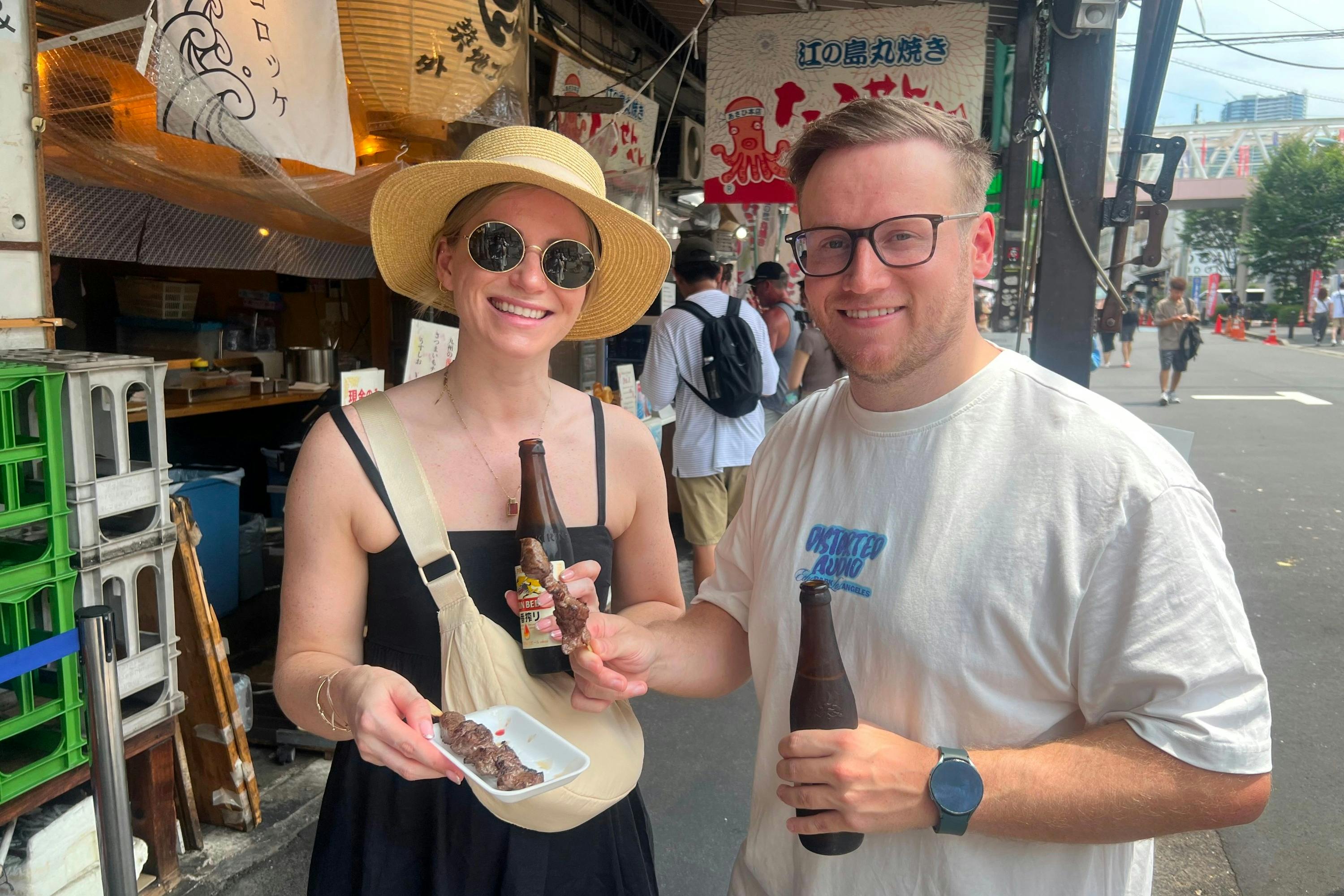 Two people smiling while holding skewered food and drinks in a lively outdoor market area.