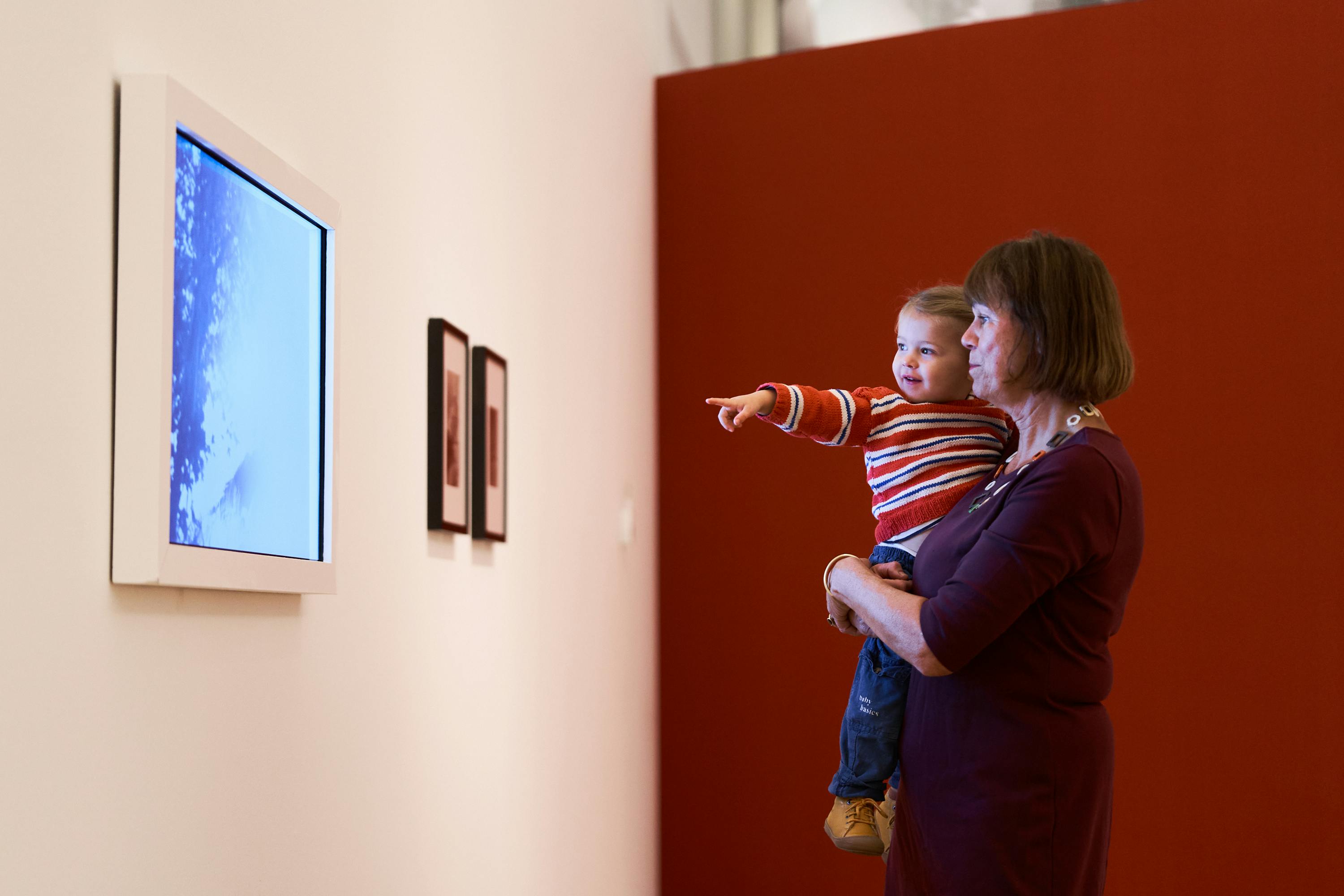 Une femme tient dans ses bras un enfant qui lui montre des photos encadrées sur un mur, tous deux regardant dans la même direction.