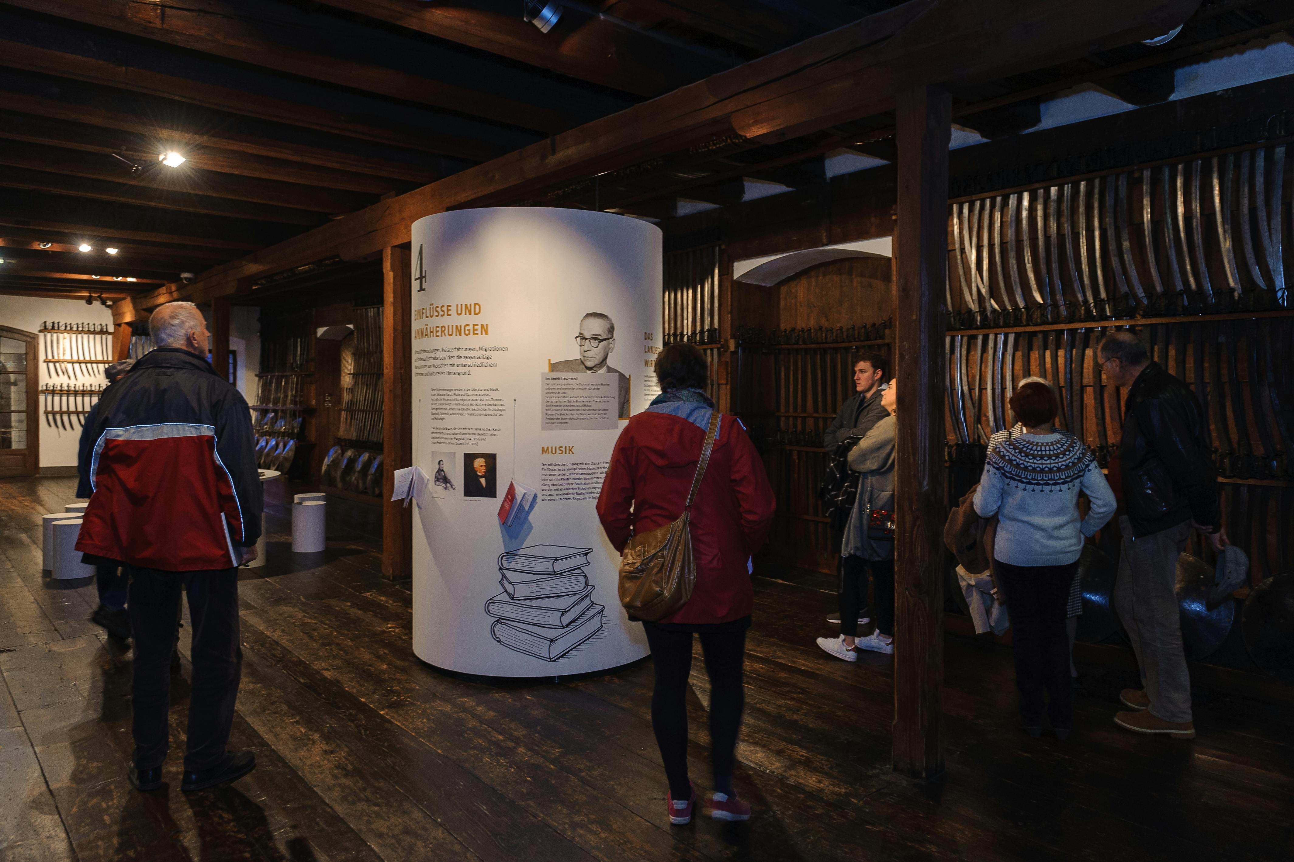 People inside a dimly lit museum, standing around a large informational display with text and images, wooden walls, and beams.