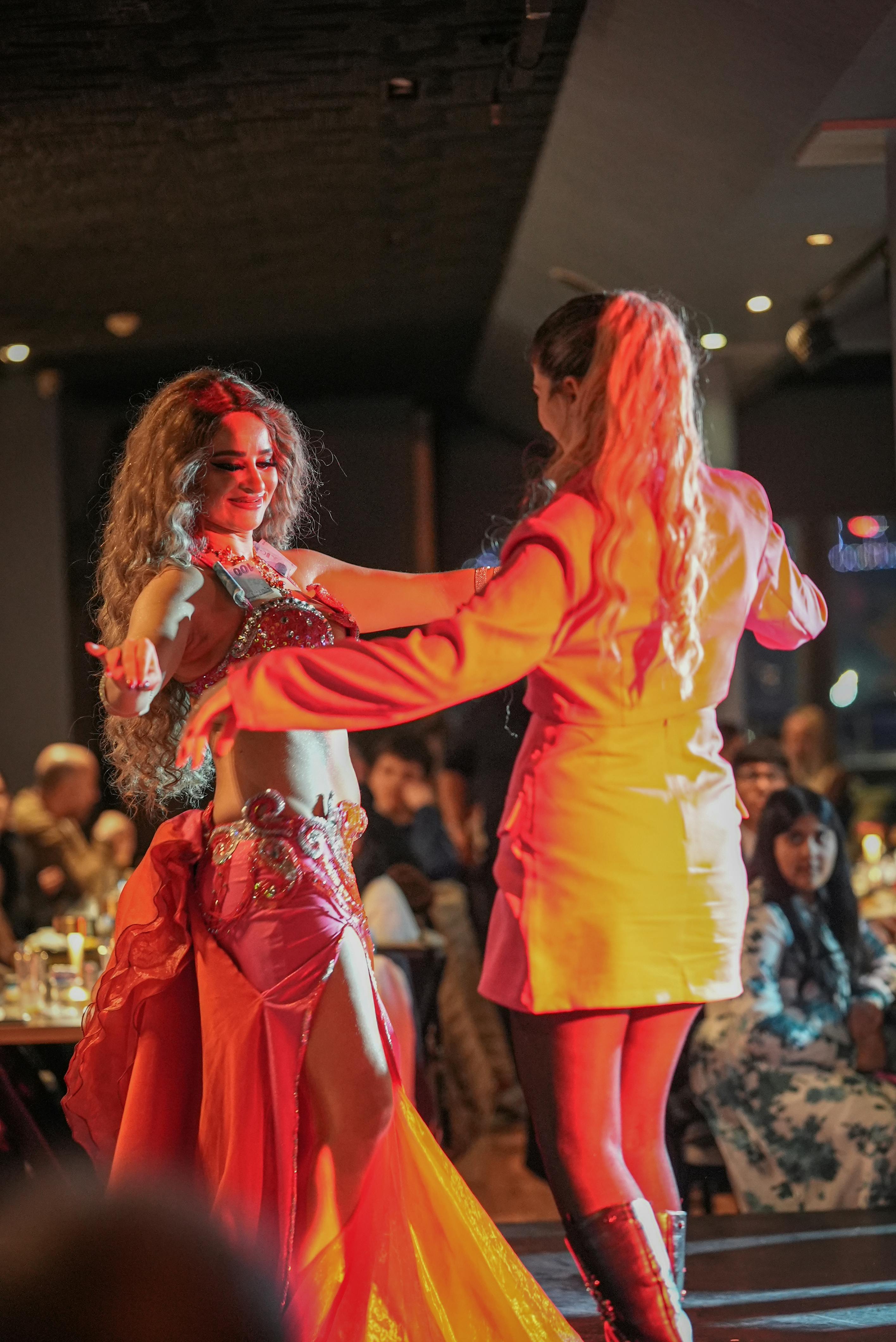 混雑した薄暗いレストランで、赤いコスチュームを着たベリーダンサーが観客と共演します。
