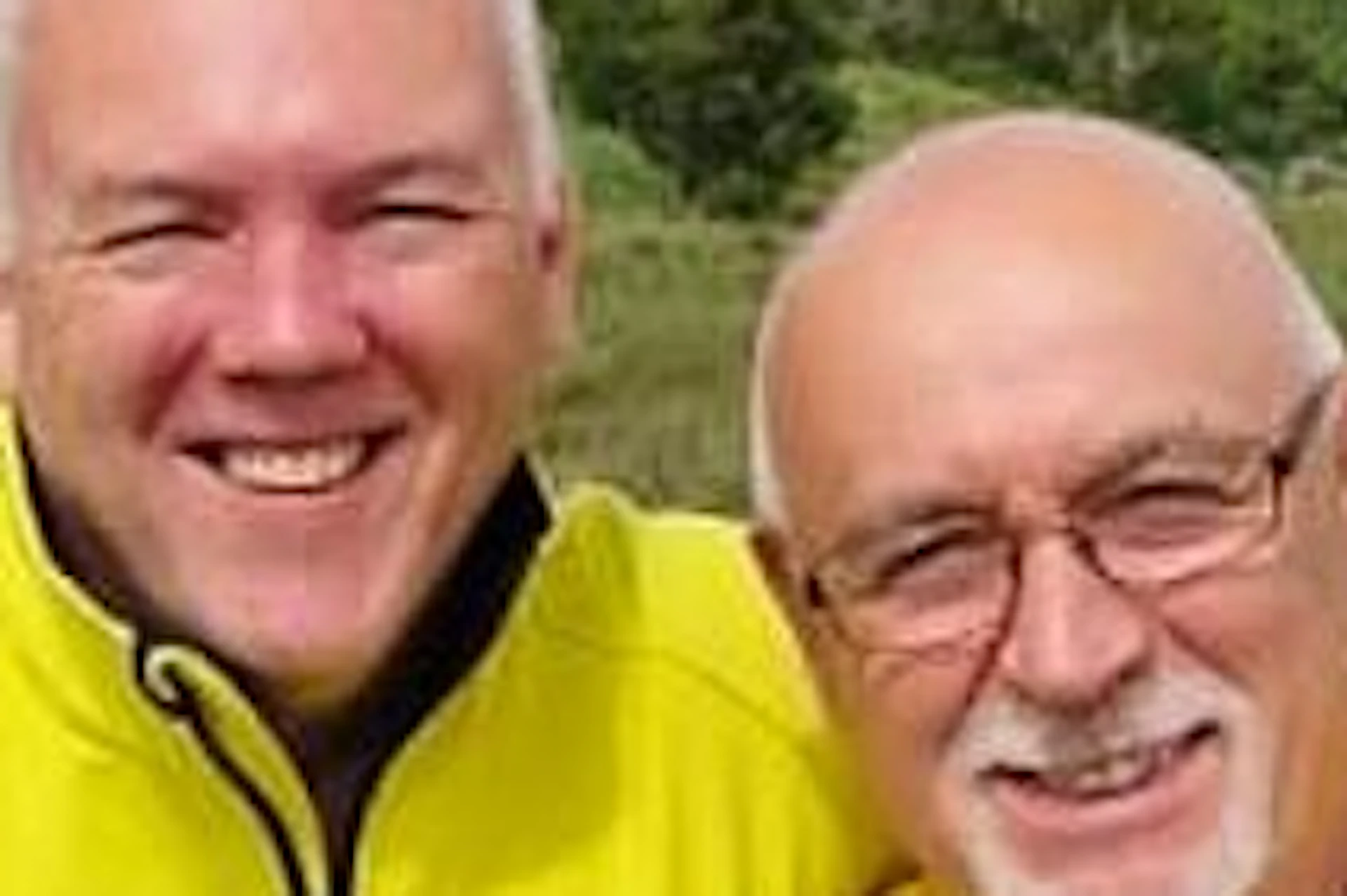 Two people wearing yellow tops smiling while standing in a scenic area with green hills and a lake in the background.