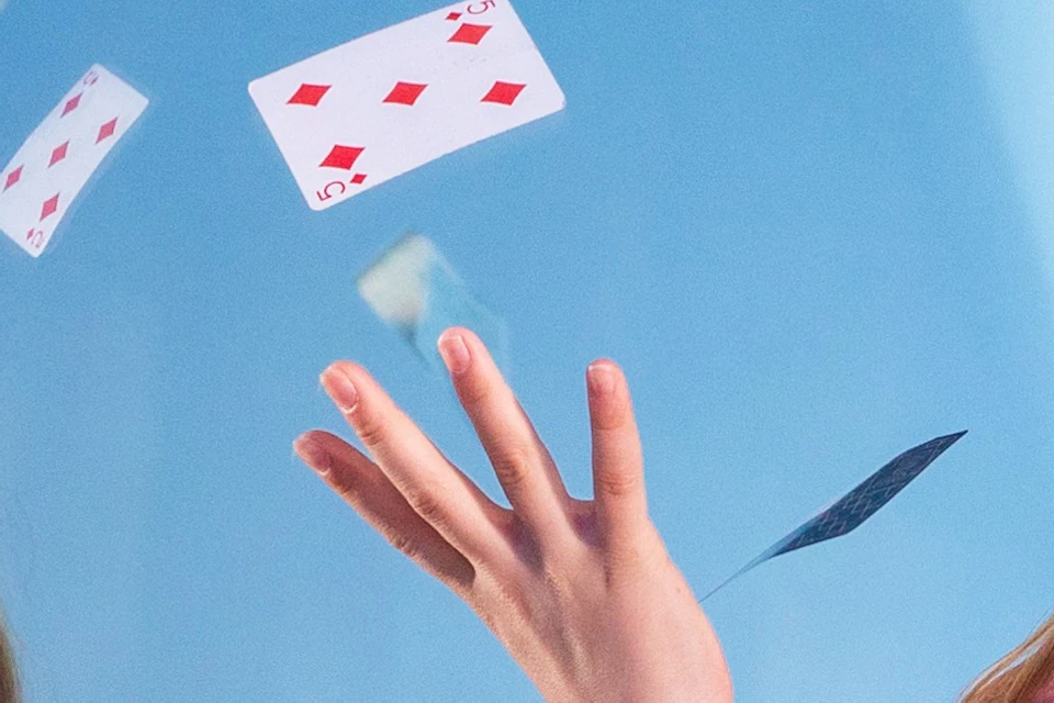 Young girl in pink shirt and jeans, juggling playing cards in front of a mirror with a modern circular light fixture above.