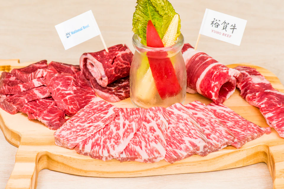 A wooden platter with slices of marbled beef, rolled beef slices, and a small jar containing vegetables and lettuce.