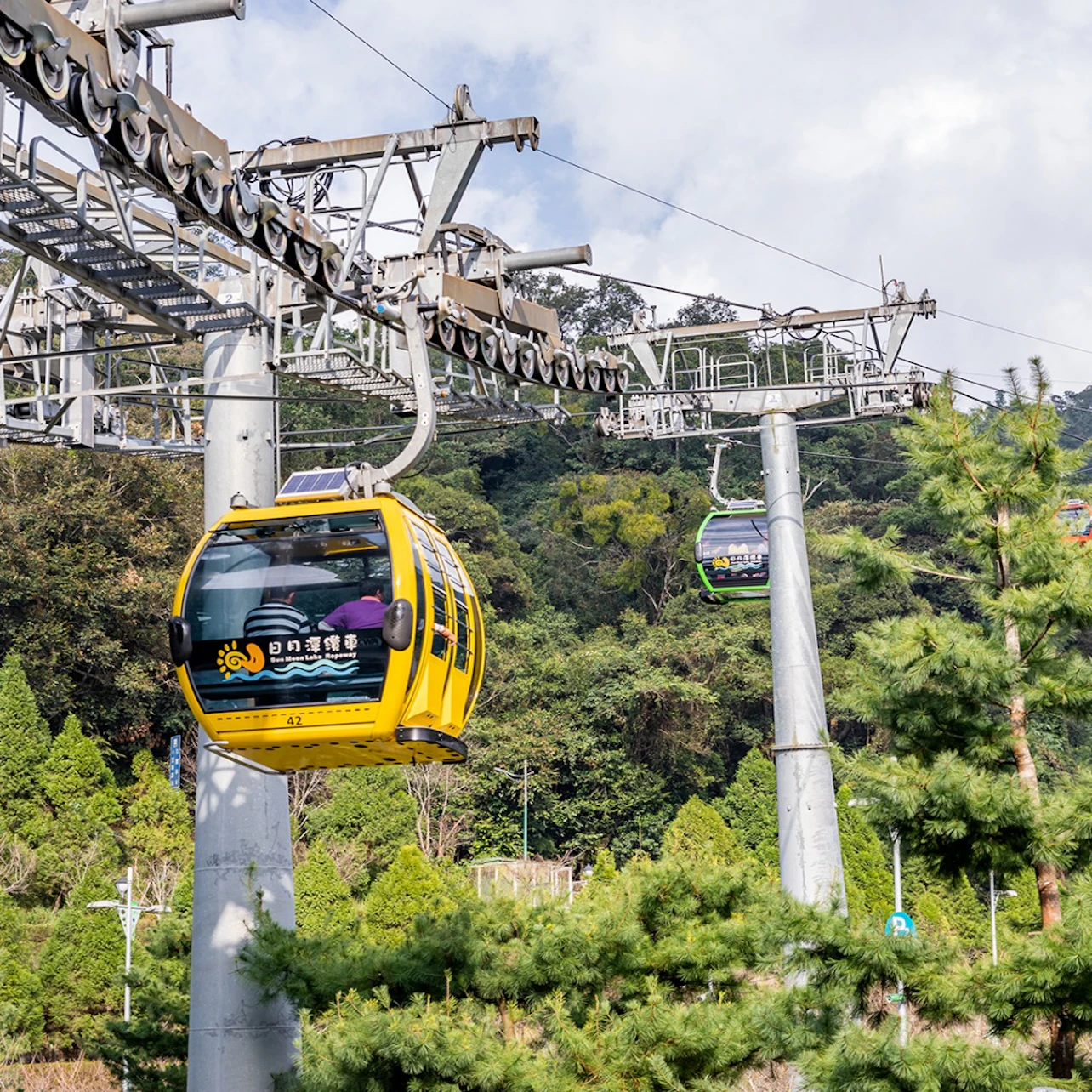 A yellow cable car on a lift system passes through a forested area under a partly cloudy sky, with another car in the background.