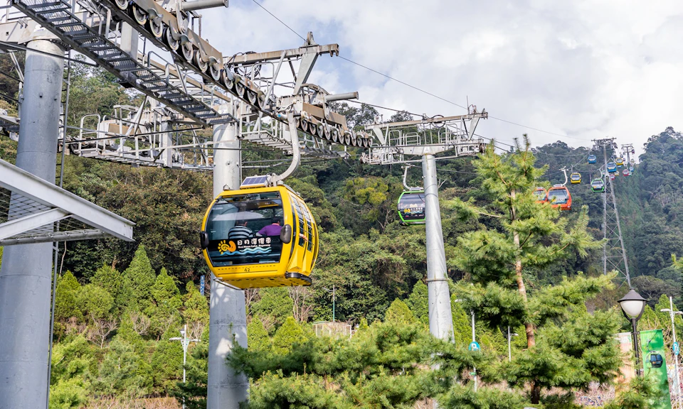 Two gondola cable cars, one yellow and one green, travel on a cable system above a forested landscape on a sunny day.