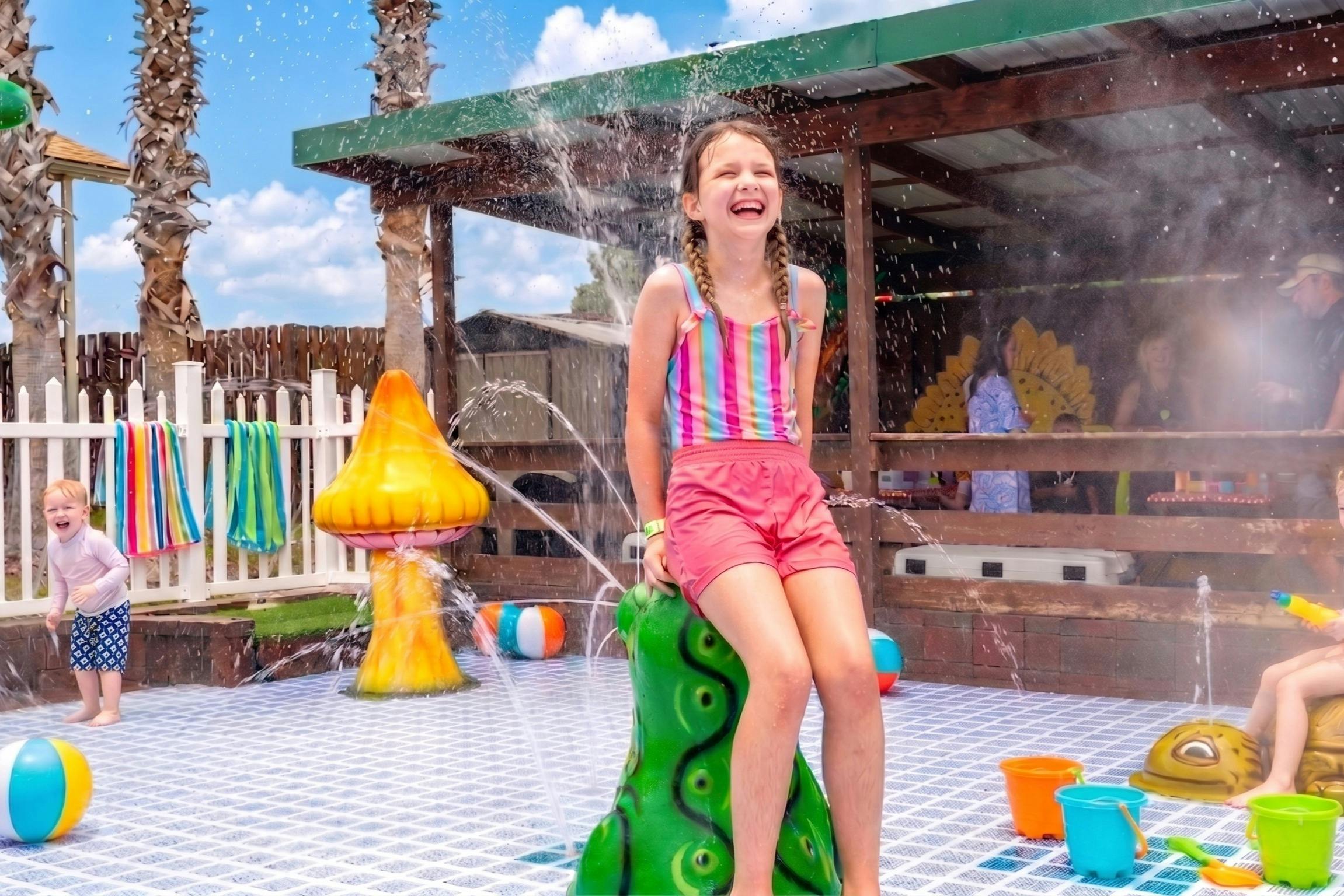 Una niña sonriente está sentada en una fuente con forma de pulpo verde en una zona de juegos con decoraciones de colores y chorros de agua.