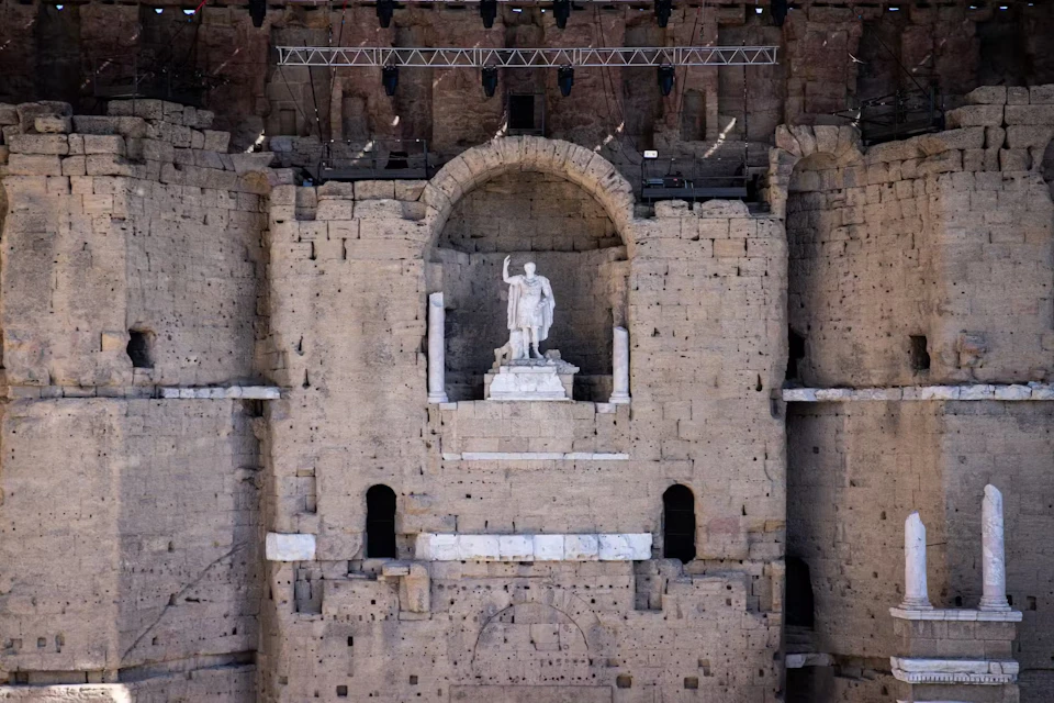 Stone wall with statue of a robed figure in an alcove. Large rectangular openings and overhead lighting fixtures are visible.