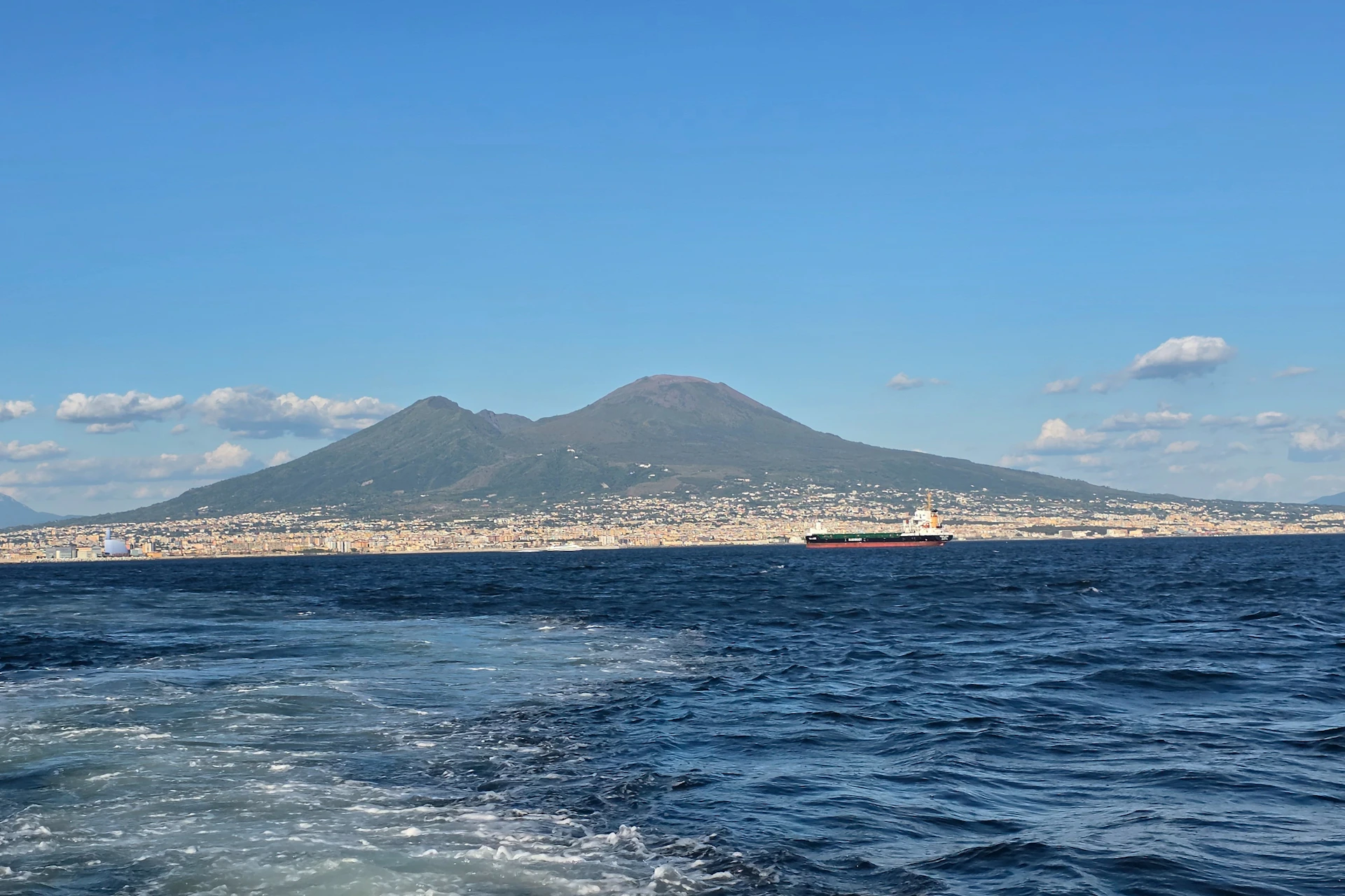 A view of Mount Vesuvius with a city in the foreground, as seen across the sea under a clear blue sky.