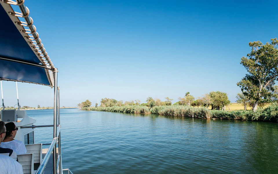 Clear blue sky over a calm river, with a boat's railing visible on the left and lush greenery along the riverbank.