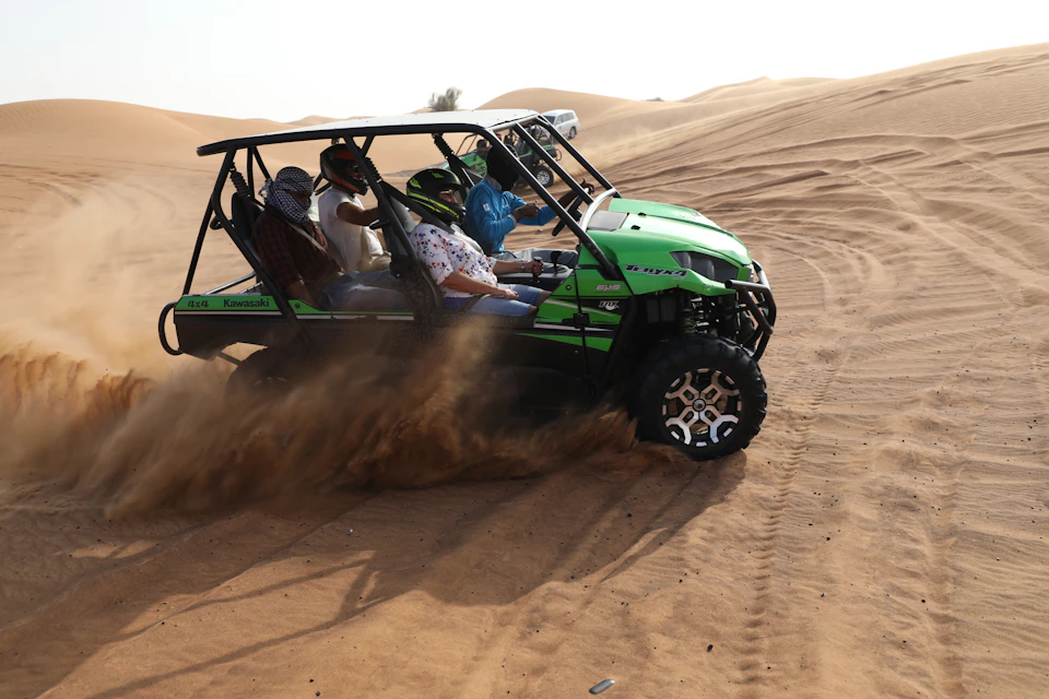 Four people riding a green off-road vehicle speeding through sandy desert dunes.