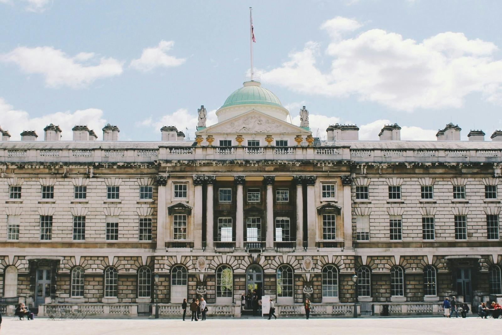 A neoclassical building with columns and a central dome, featuring a flag on top, with people in front on a sunny day.