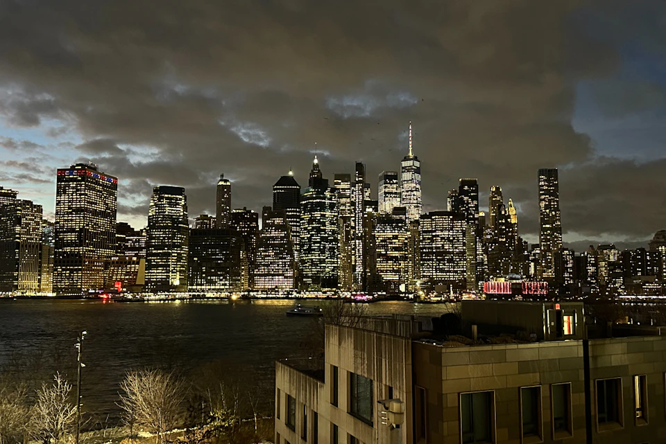 A city skyline at night with illuminated skyscrapers under a cloudy sky, reflecting on the water below.