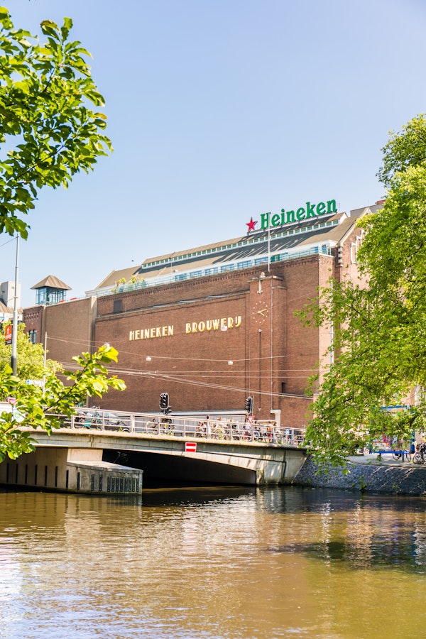 "Heineken Brouwerij" 표지판이 있는 벽돌 건물, 지붕의 Heineken 로고, 운하 위의 다리, 푸른 하늘 아래 푸른 나무.