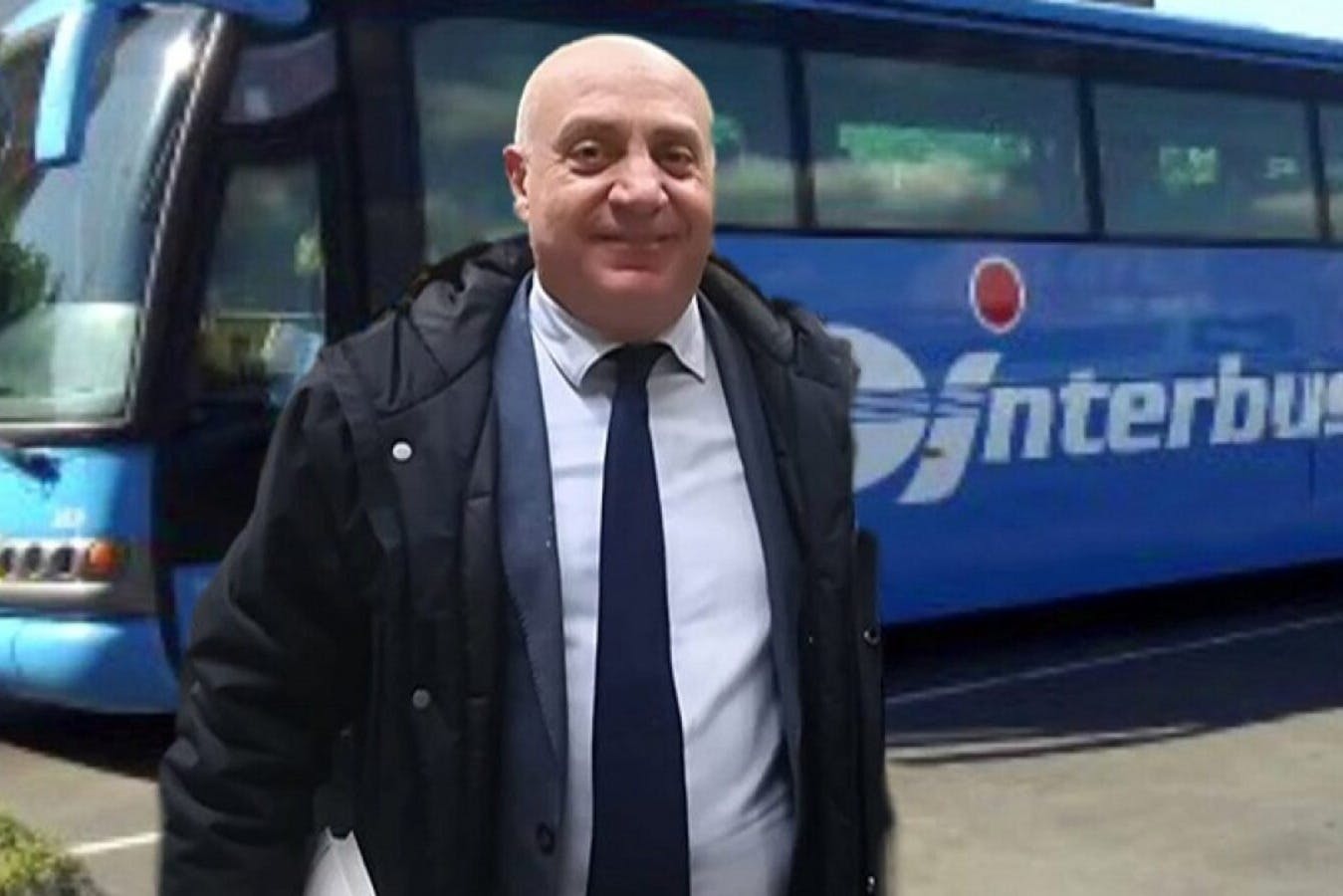 Smiling man in a dark jacket and tie stands in front of a blue bus.