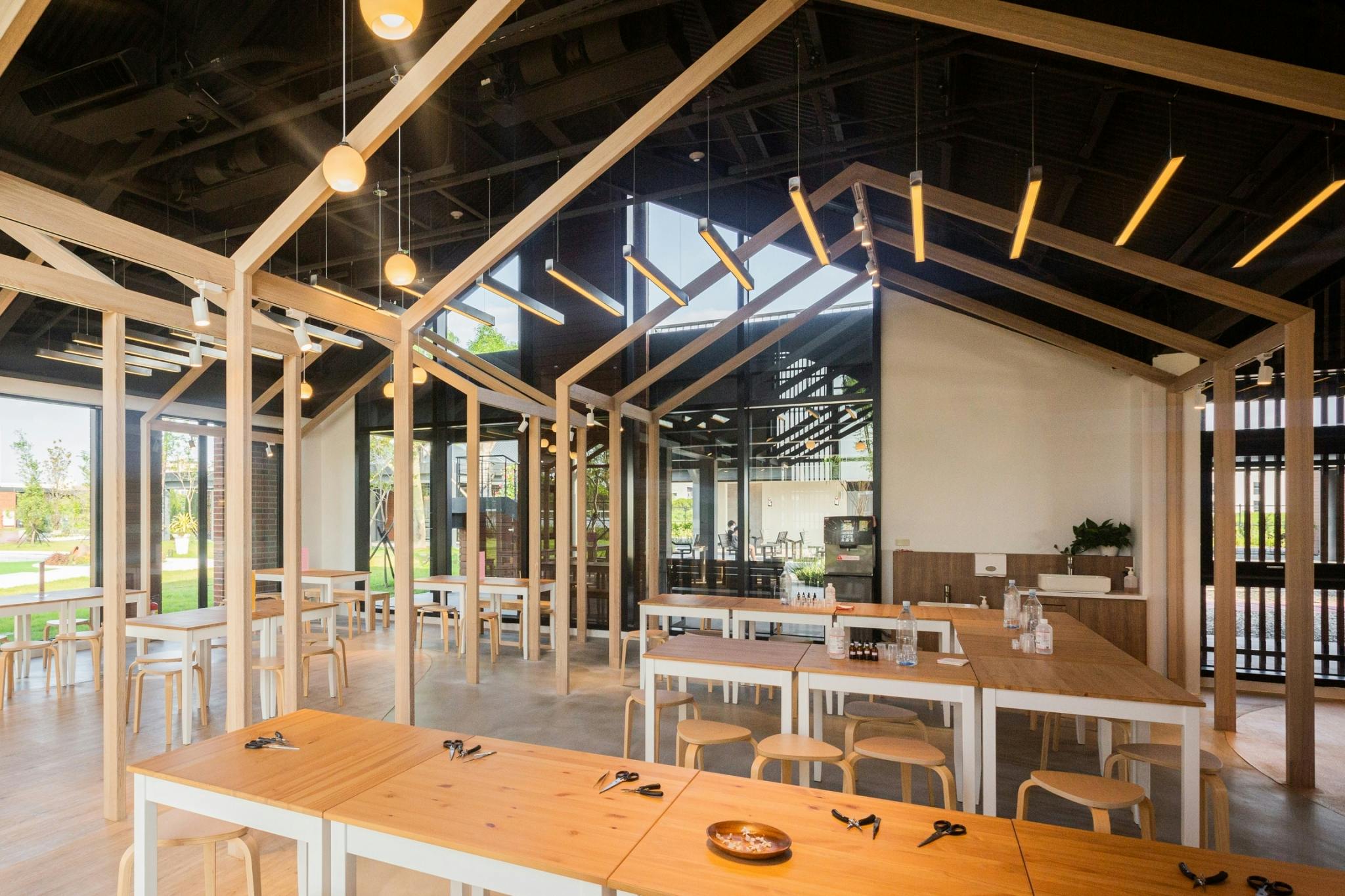 A modern dining area with wooden tables, stools, and decorative lighting under a high ceiling with large windows.