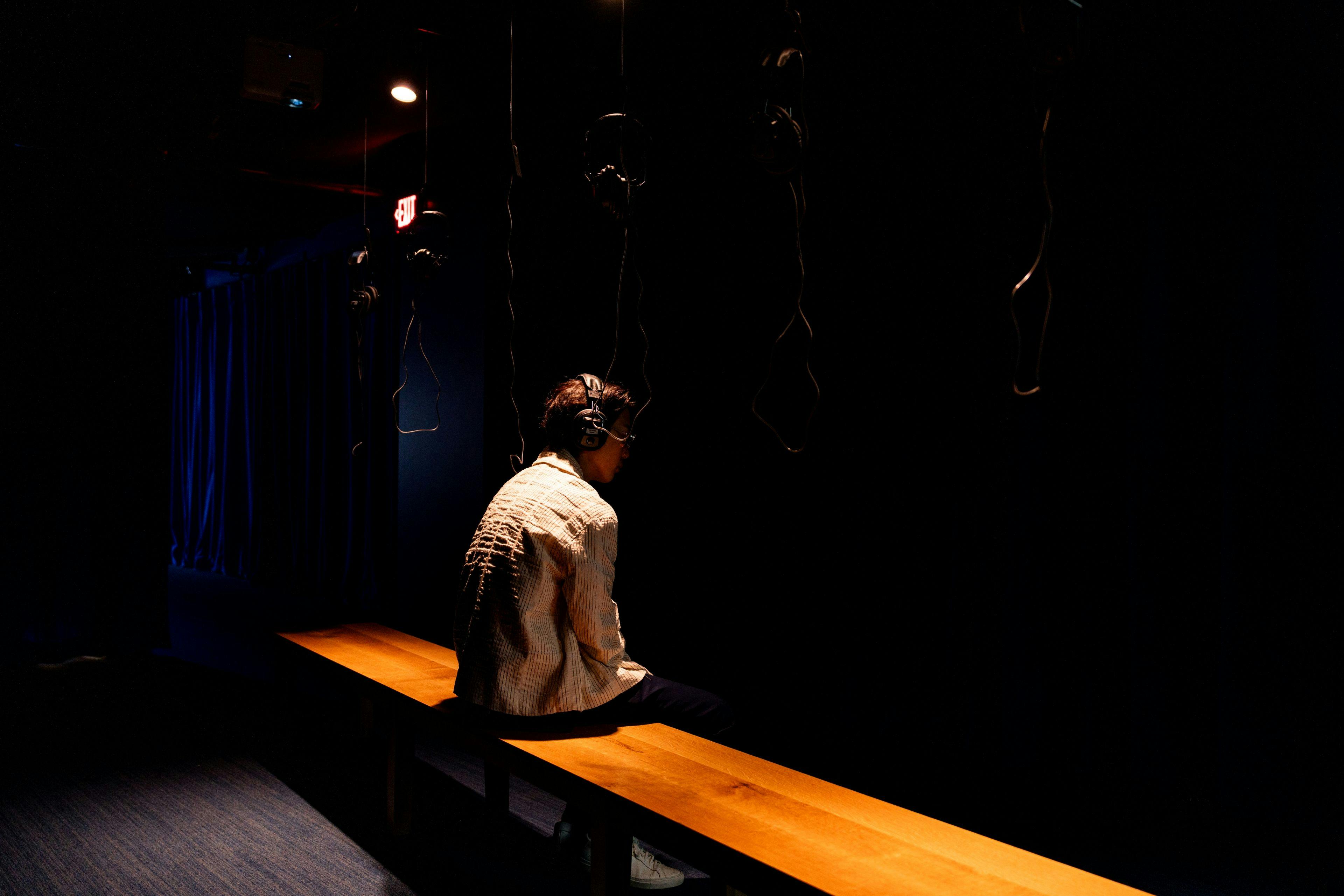 Eine Person mit Kopfhörern sitzt allein auf einer Holzbank in einem schwach beleuchteten Raum mit hängenden Kabeln.