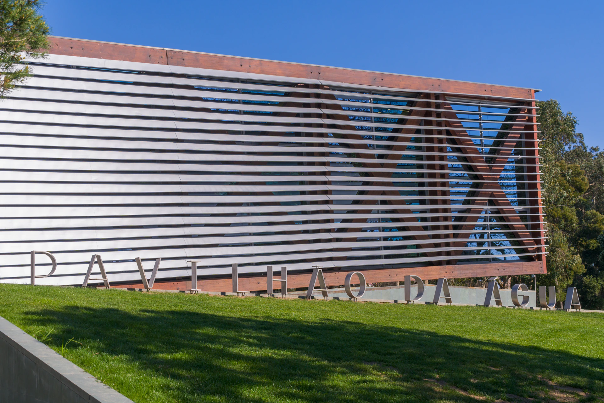 Modern building with horizontal slats, "PAVILHAO DA" partially visible sign, set against a clear blue sky with green grass in front.