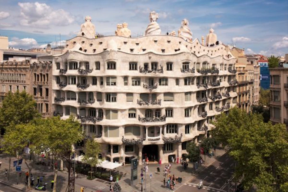 Multi-story, wavy building with ornate balconies and sculptures on the rooftop, surrounded by trees and pedestrians.