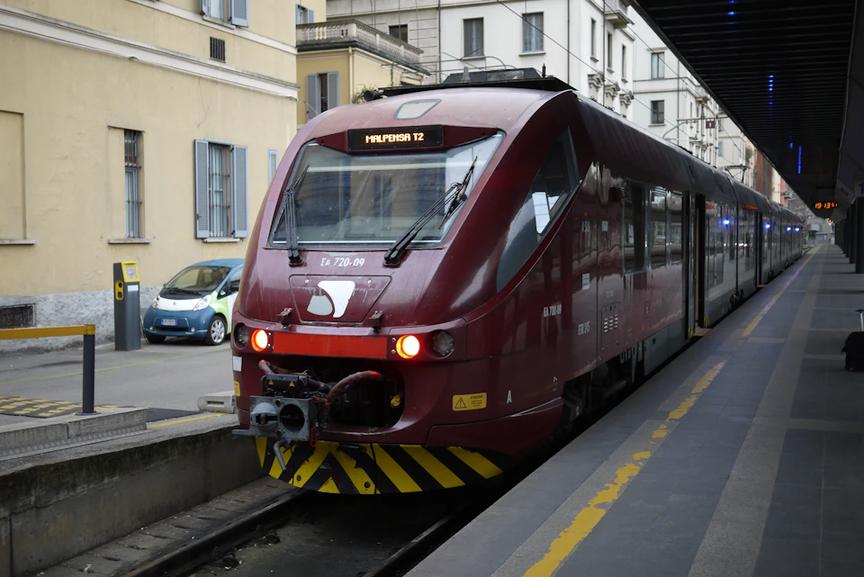 A red train marked "Malpensa T2" at a station platform, with buildings and parked cars in the background.