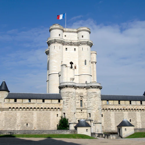 Ett högt, historiskt stenslott med torn och en fransk flagga på toppen, under en blå himmel med spridda moln.