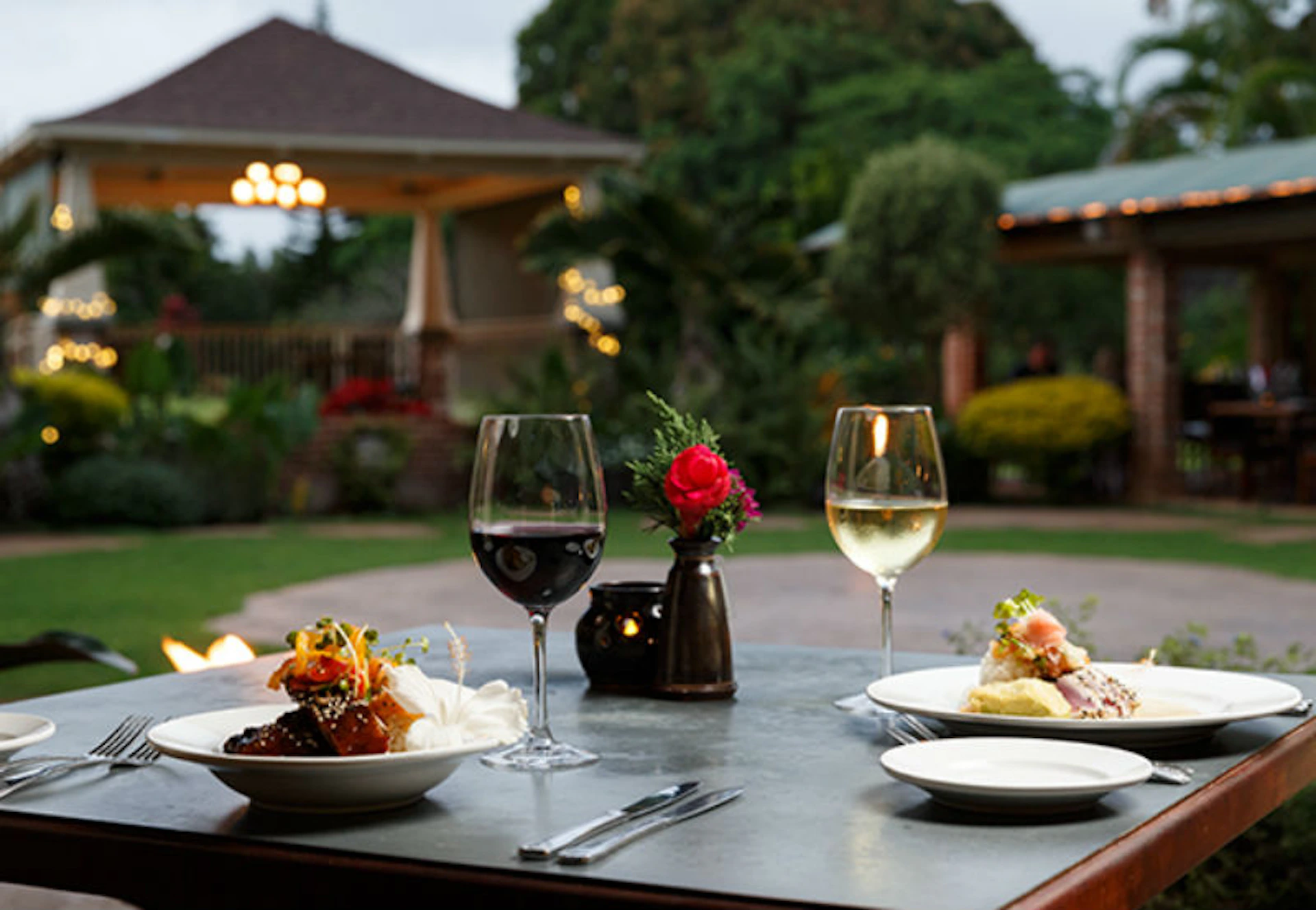 Two plates of food and two glasses of wine on an outdoor dining table, with a central flower vase and garden in the background.