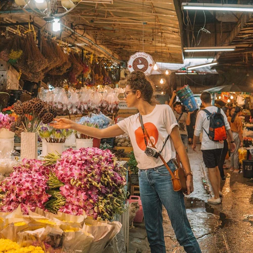 Eine Person wählt Blumen in einer belebten Markthalle aus, in der verschiedene Blumen in Sträußen ausgestellt sind und andere Kunden stöbern.