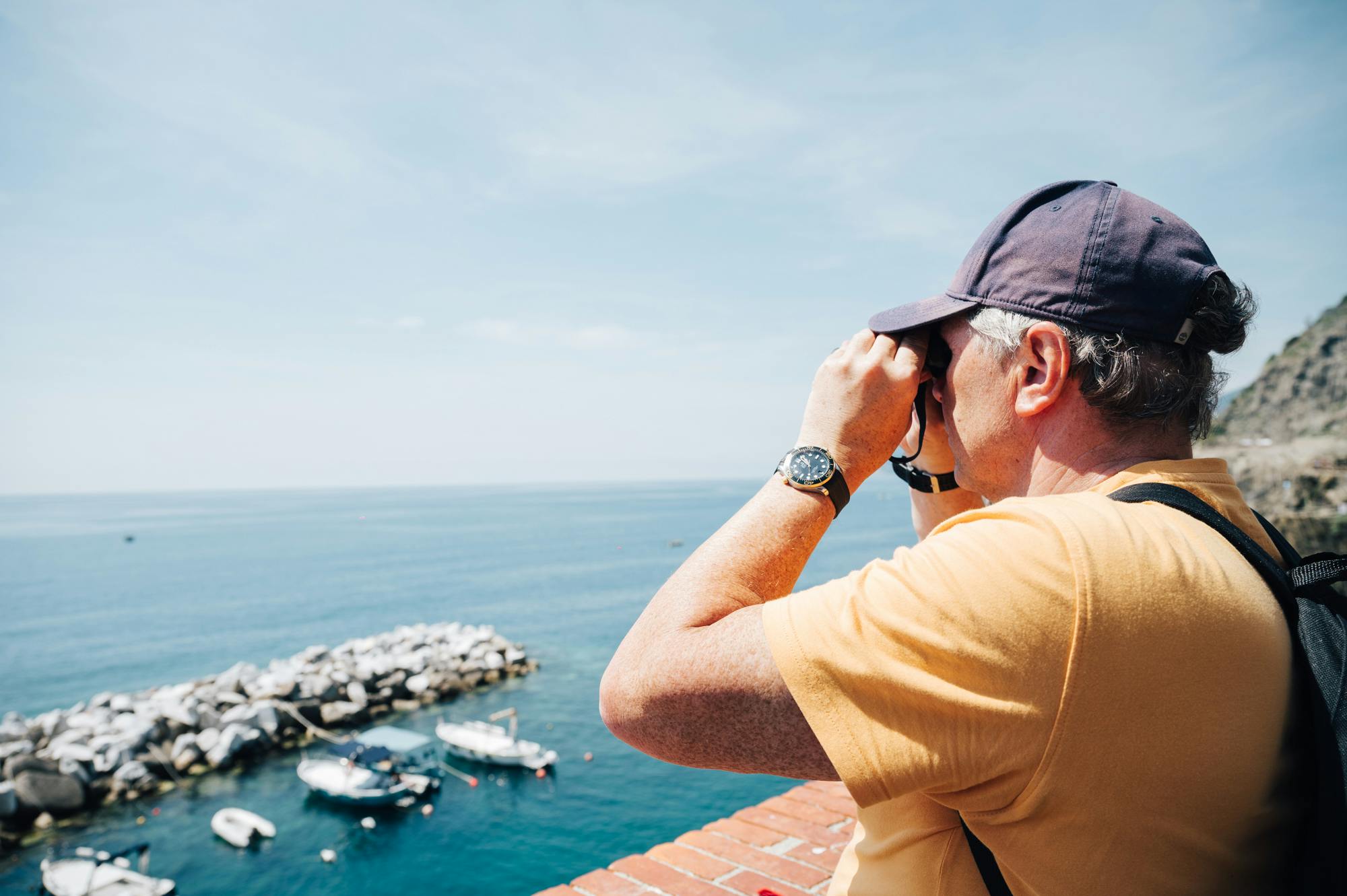穿着黄色衬衫和帽子的人使用双筒望远镜，从带有砖栏杆的阳台上眺望大海。