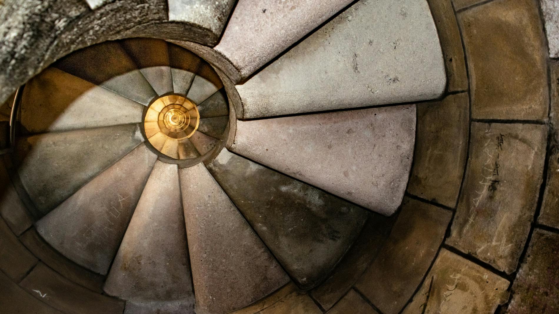 Spiral stone staircase with worn steps, viewed from above with a yellow light at the center illuminating the stairs.
