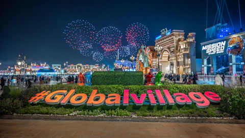 Night scene of #GlobalVillage with illuminated attractions, crowds of people, colorful fireworks, and a sign for "Mission Speed Stunt Show."