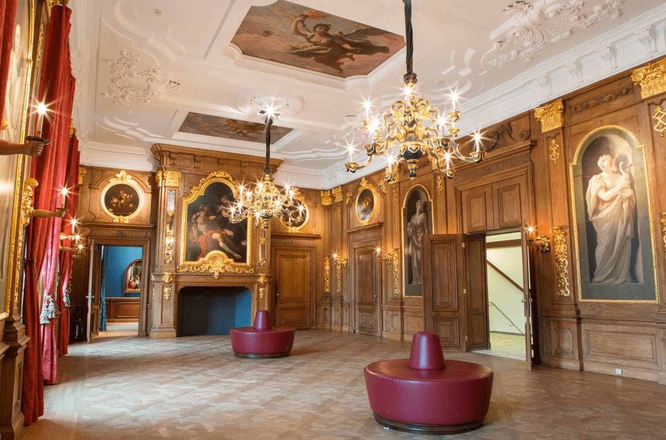 Περίτεχνο δωμάτιο με πολυελαίους, πίνακες ζωγραφικής σε τοίχους και οροφή, σκαλιστούς ξύλινους τοίχους και στρογγυλά κόκκινα καθίσματα.