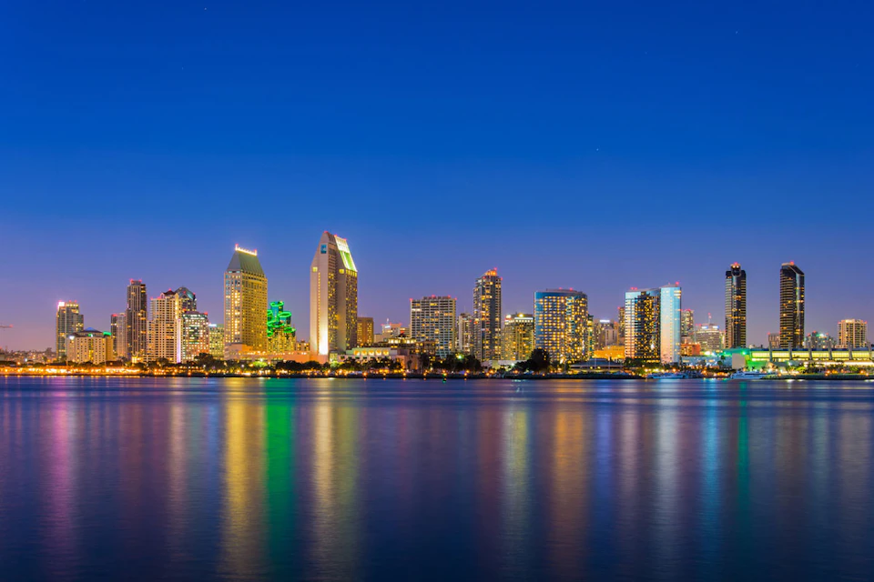 City skyline with illuminated buildings reflected on calm water, under a clear, deep blue evening sky.