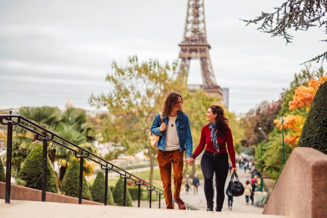 一对夫妇在埃菲尔铁塔（ Eiffel Tower ）附近的户外手拉手散步，在多云的日子里，后面有树木和人。