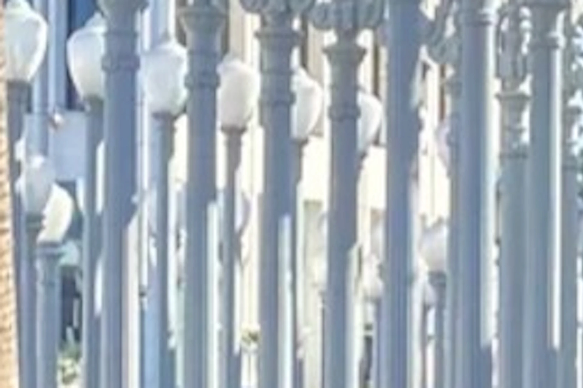 Two people stand near a large outdoor installation of numerous street lamps. A tall building and palm trees are in the background.