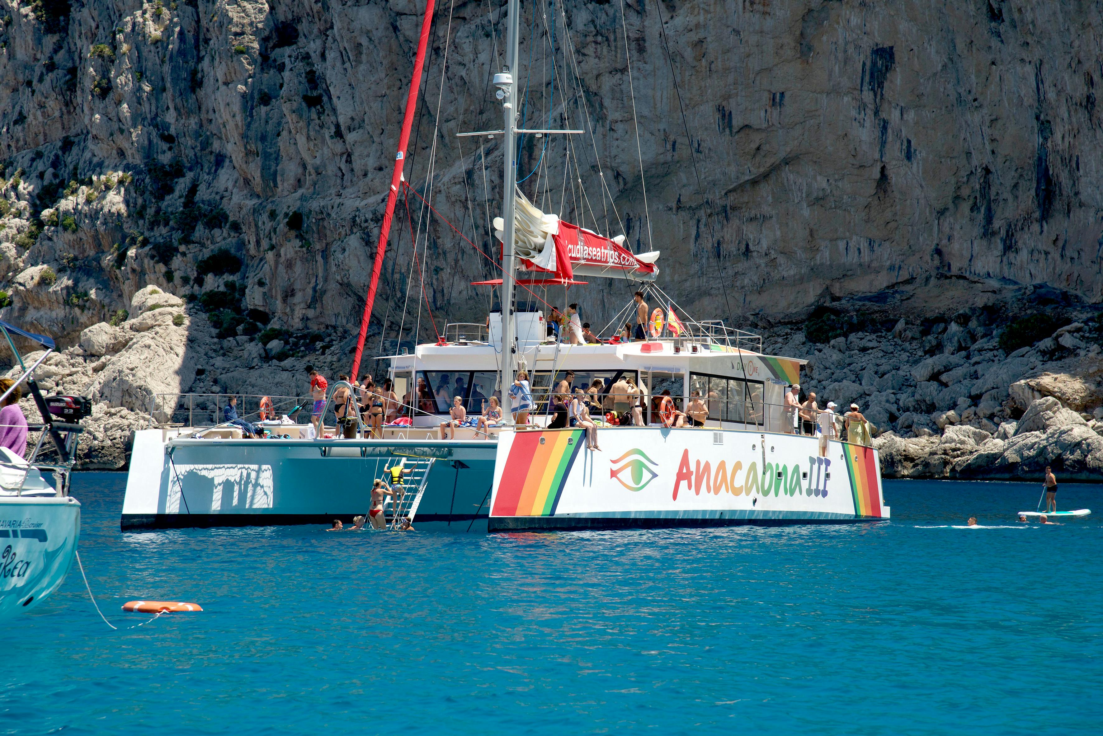 Un catamaran nommé "Anaconda III" avec des personnes à bord est ancré près d'une falaise rocheuse dans une eau bleue claire.