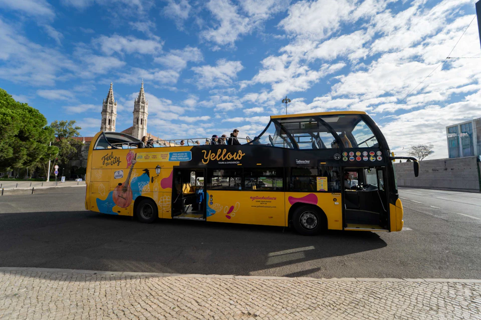 A yellow double-decker tour bus with an open upper deck, parked on a street near a building with tall spires under a partly cloudy sky.