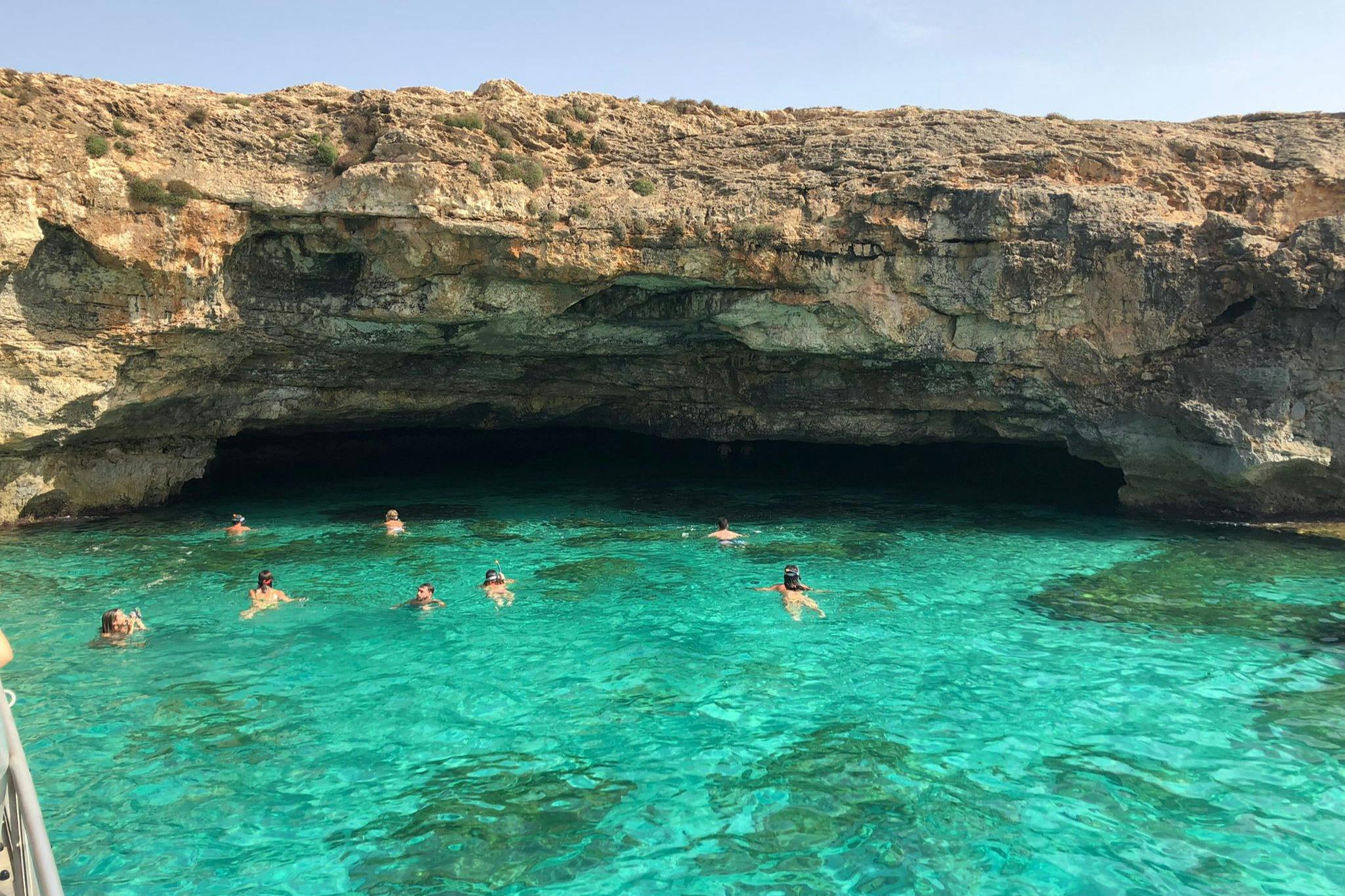 Άνθρωποι που κολυμπούν σε καθαρά γαλαζοπράσινα νερά κοντά σε μια βραχώδη σπηλιά κάτω από έναν ηλιόλουστο ουρανό.