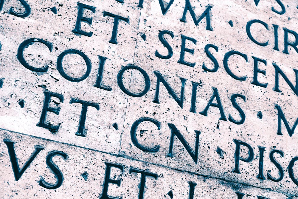 Close-up of ancient Roman-style engraved text on a weathered stone surface, with visible letters including "COLONIAS."