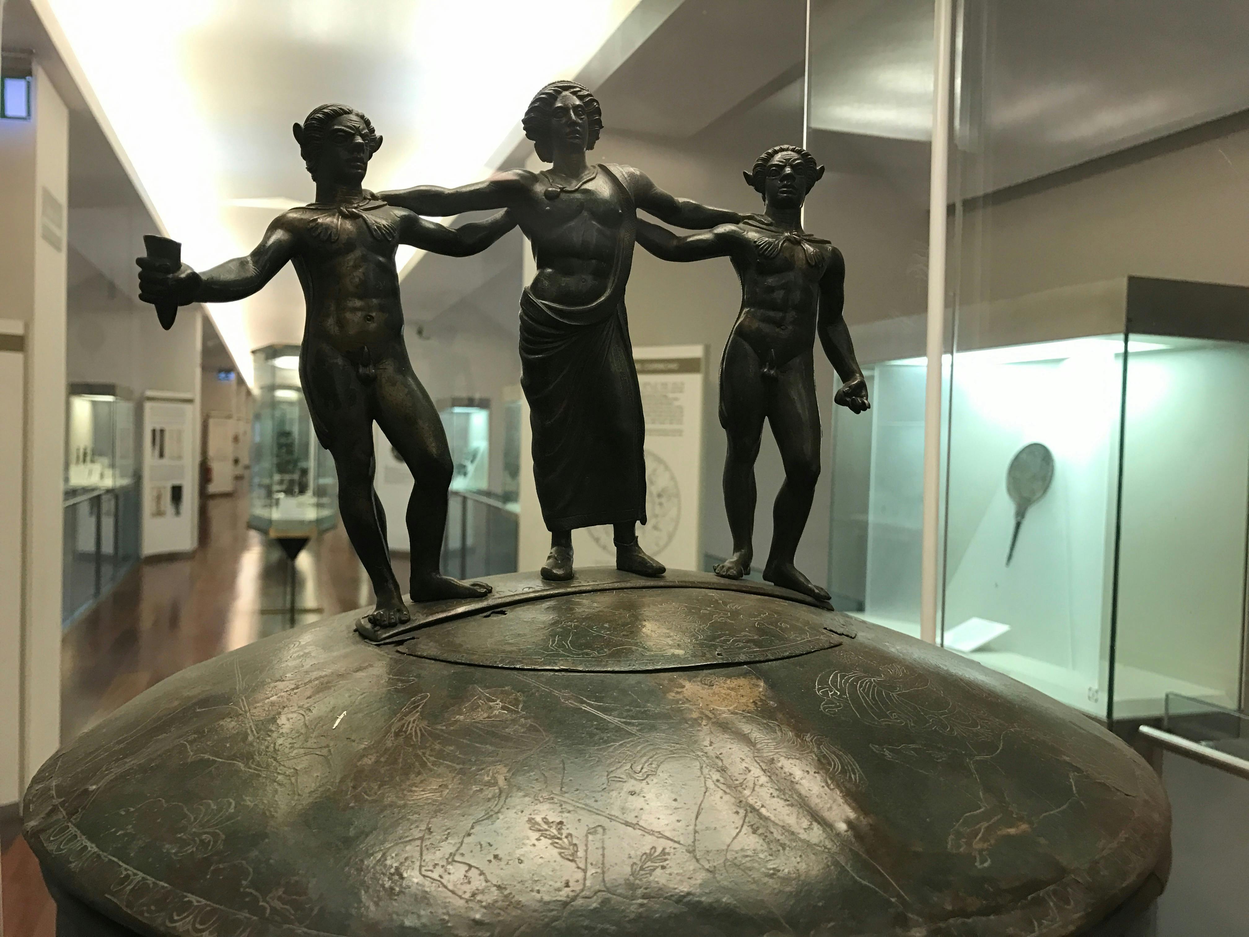 Een bronzen sculptuur met drie menselijke figuren op een versierde sokkel, in een vitrine van een museum.