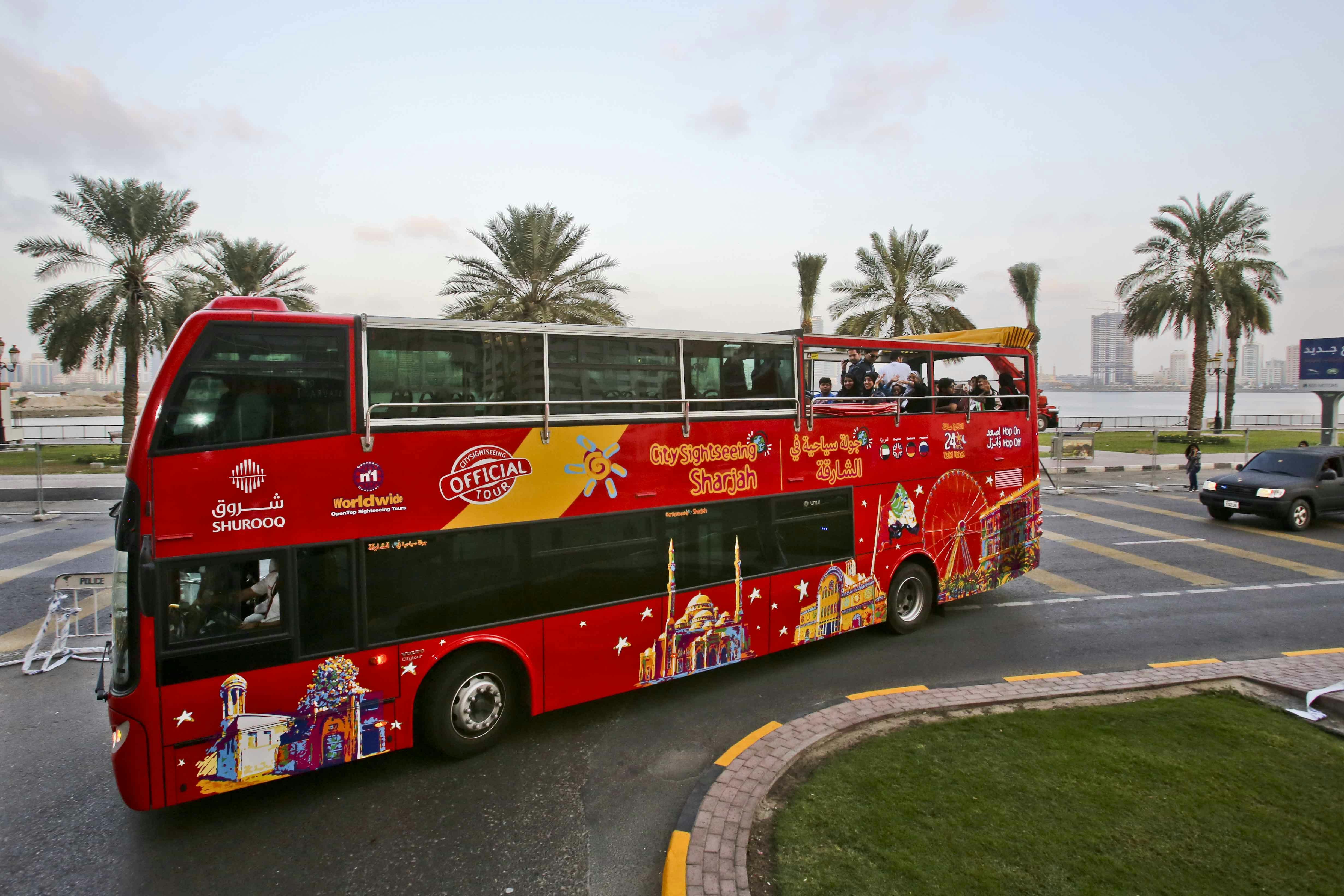 A red double-decker tour bus with “City Sightseeing Sharjah” branding. Passengers are on the open-top upper deck. Palm trees in the background.