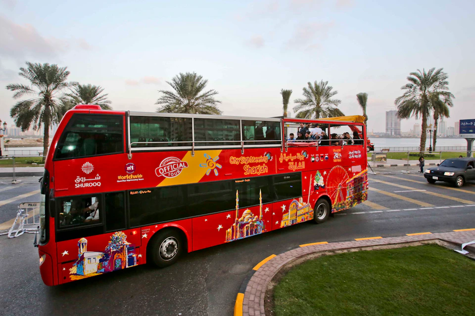 A red double-decker tour bus with “City Sightseeing Sharjah” branding. Passengers are on the open-top upper deck. Palm trees in the background.