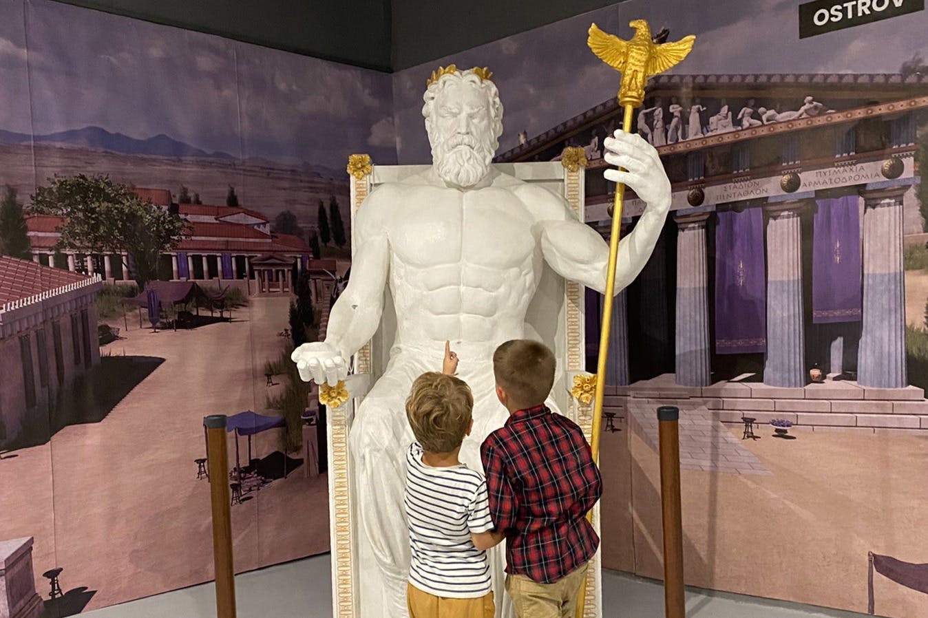 Two children stand before a large statue of a seated, bearded figure, pointing at it. A mural of an ancient building is in the background.