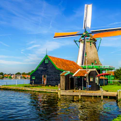 En traditionell väderkvarn och trähus med orange tak bredvid en flod, under en klarblå himmel.