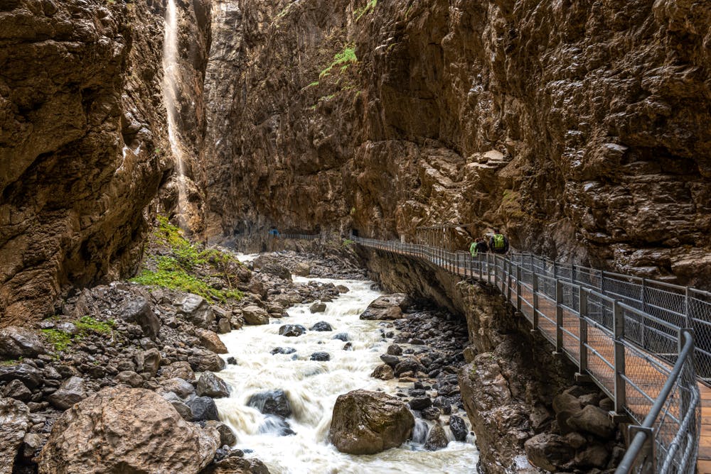 Narrow canyon with a rushing river, hiking pathway with railings on the right, and a small waterfall on the left.