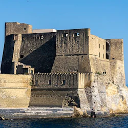 Forntida stenfästning med fyrkantiga torn på en stenig strand vid havet, under en klarblå himmel.
