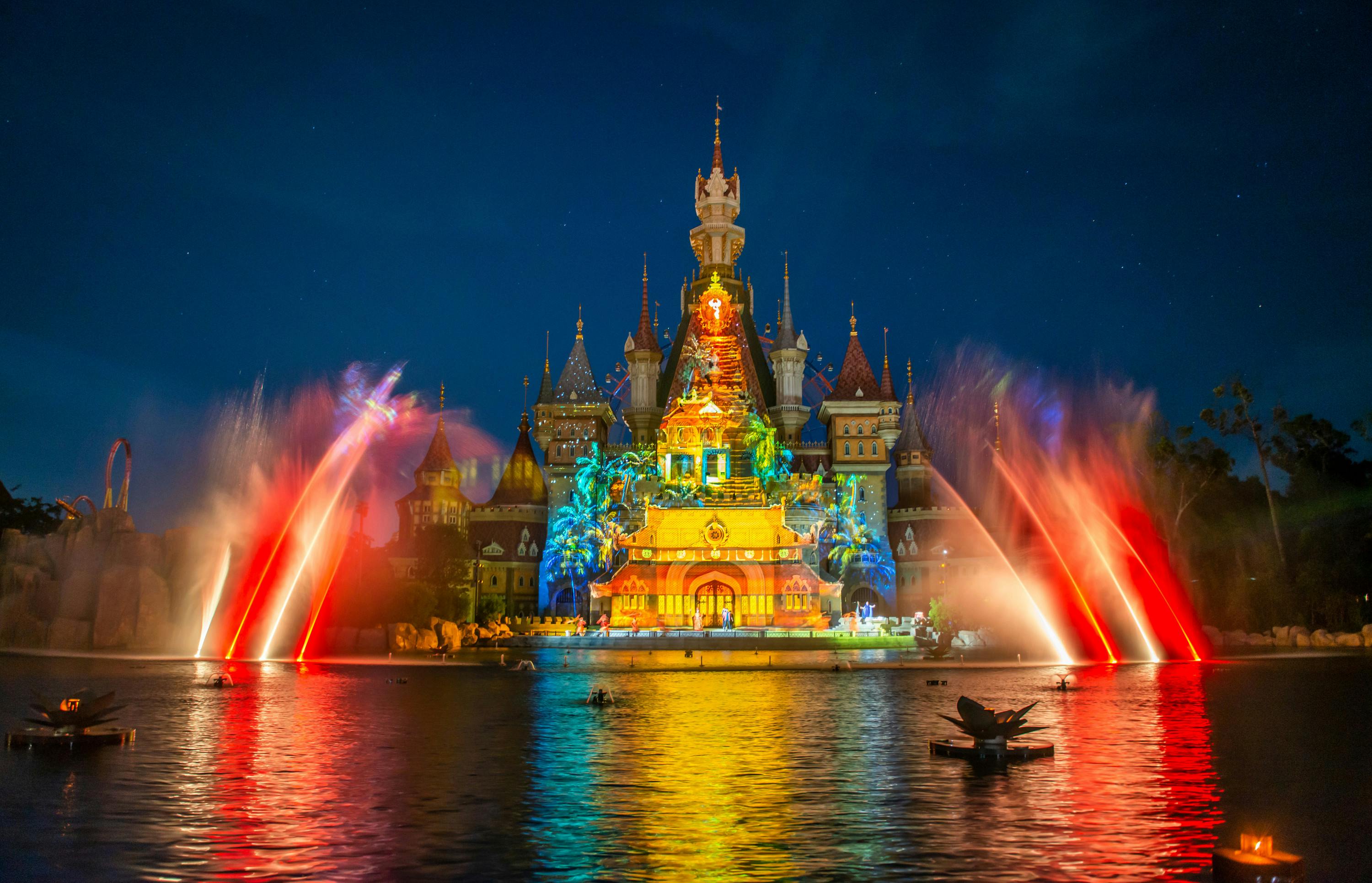 A brightly illuminated castle at night with colorful water fountains and light projections in front of it.