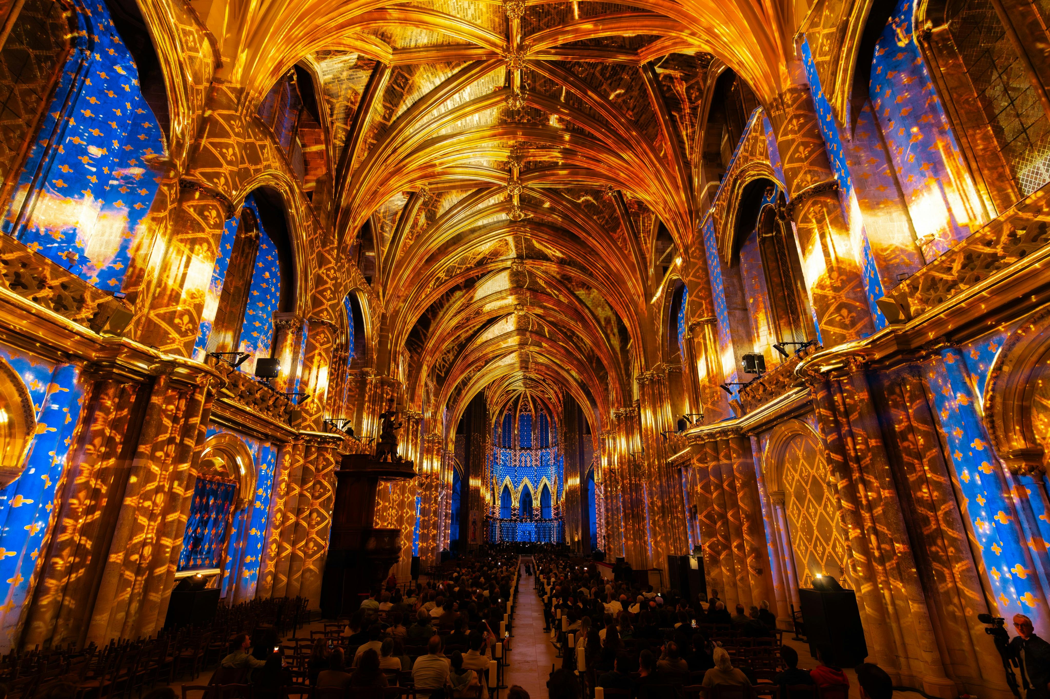 An ornate cathedral interior illuminated with vibrant golden and blue lighting, with people seated in rows observing the scene.