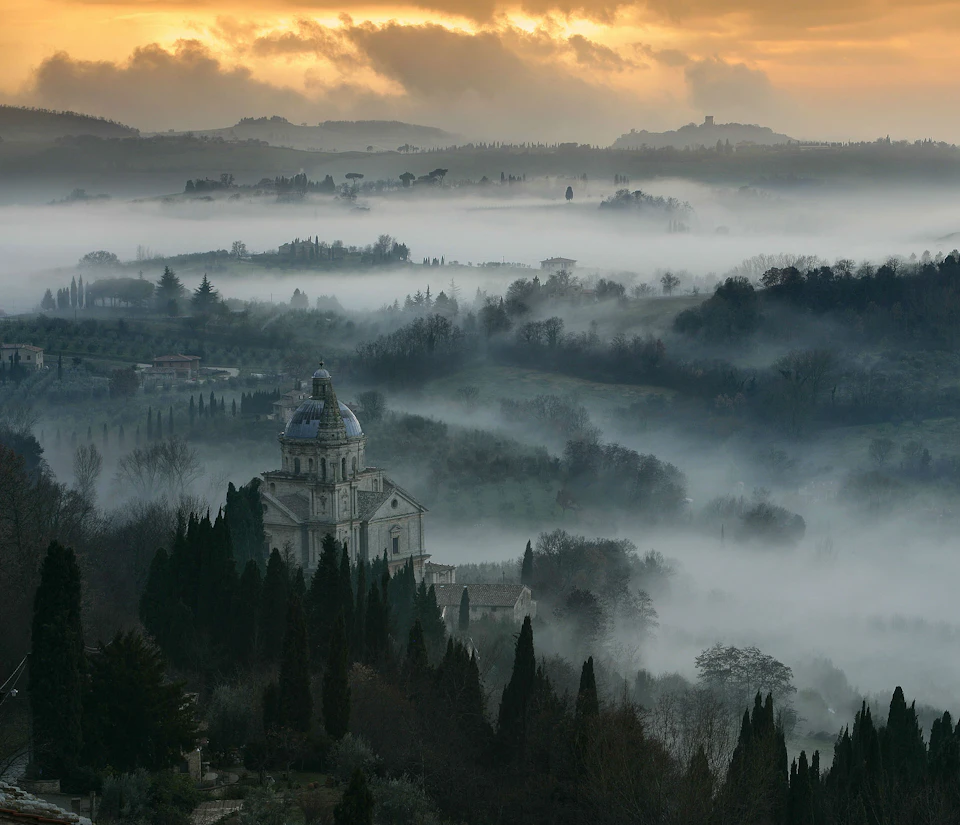 A church surrounded by cypress trees is set in a misty, rolling landscape at dawn with a rising orange and gray sky.