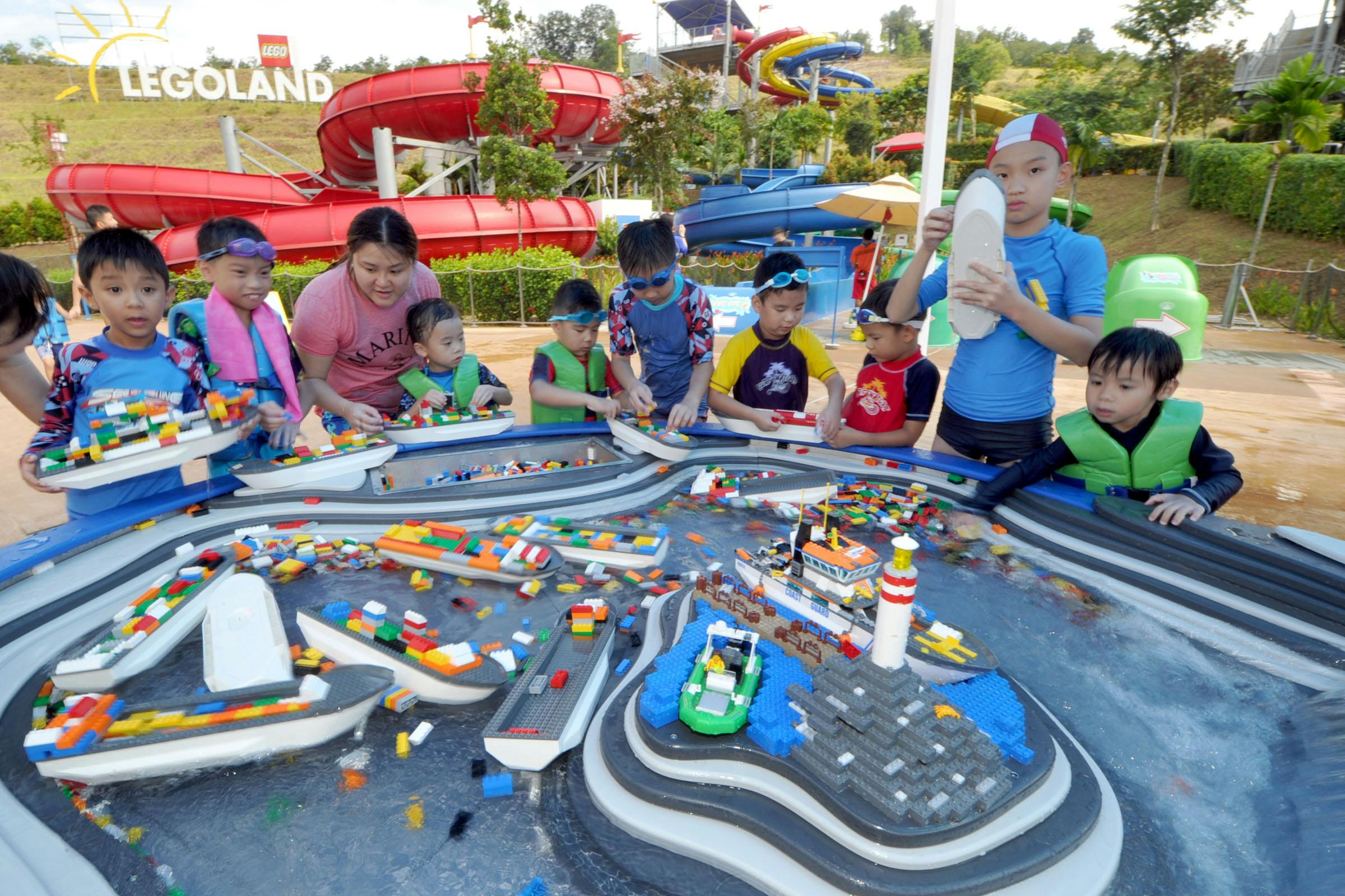 Children and an adult play with floating LEGO bricks in a water-themed play area at LEGOLAND, with large water slides in the background.