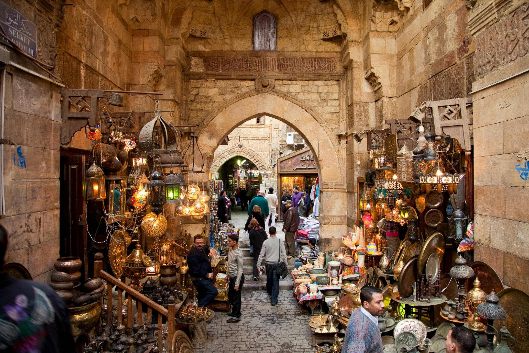 A bustling marketplace with ornate metalware and lamps on display, surrounded by people and ancient archways in a narrow alley.